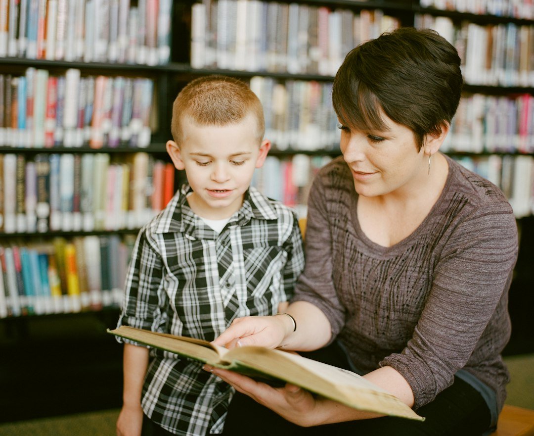 Mujer y niño pequeño leyendo juntos un libro abierto en una biblioteca con estantes de fondo